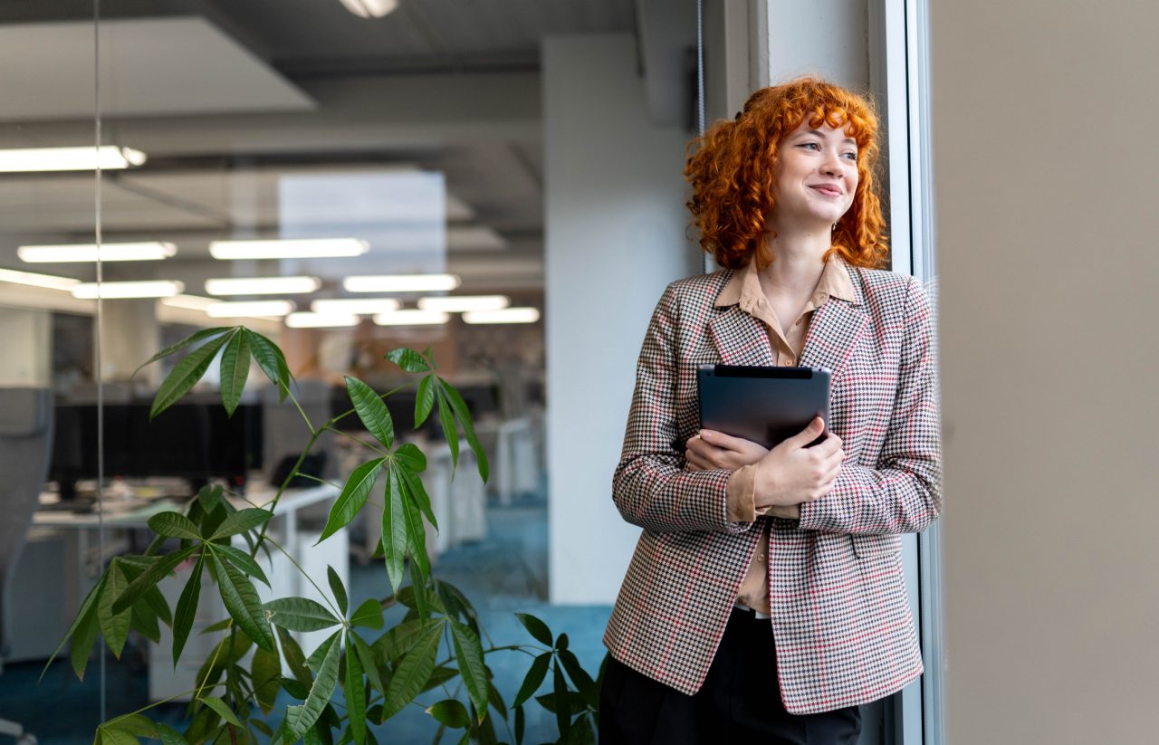 Smiling young redhead intern looks out large glass window.