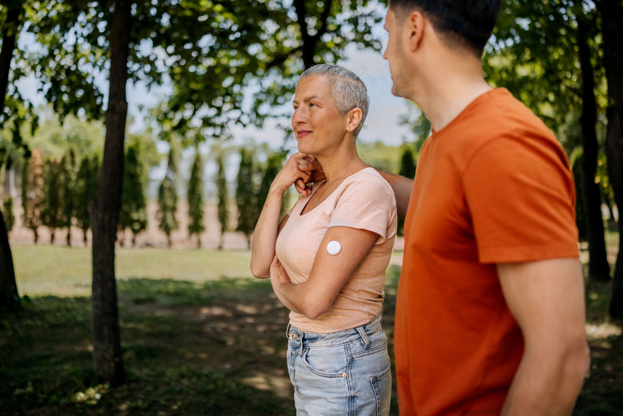 A couple walking together in a park, with the woman wearing a diabetic sensor on her upper arm. The scene reflects health awareness, daily activity, and supportive partnership