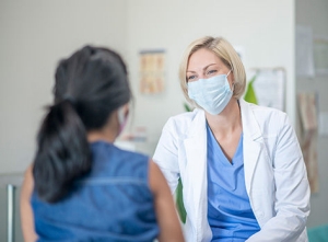 a female patient speaking with her physician in a doctors office, both are wearing masks.