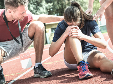 A young woman stops running when she injures her knee. Her coach attends her.