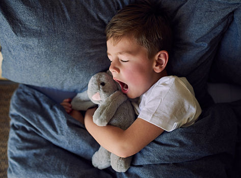 A young child lies in bed with his eyes closed.  He is yawning.