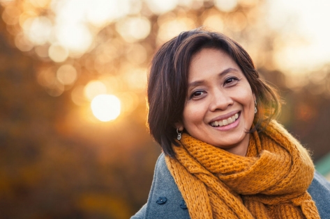 Una mujer sonriente de alrededor de 40 años al aire libre en otoño, con el sol que brilla detrás de ella.