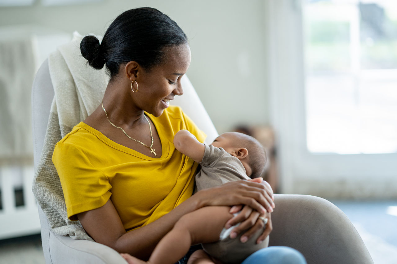 A new Mother sits in her baby's nursery with the infant in her arms and held to her chest, as she nurses him.  She is dressed casually in a yellow t-shirt and looking down at the baby with a smile.