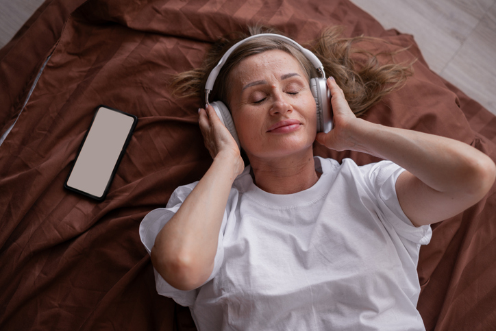 Top view on old woman listening to music in headphones on bed with smartphone near her. Copy space