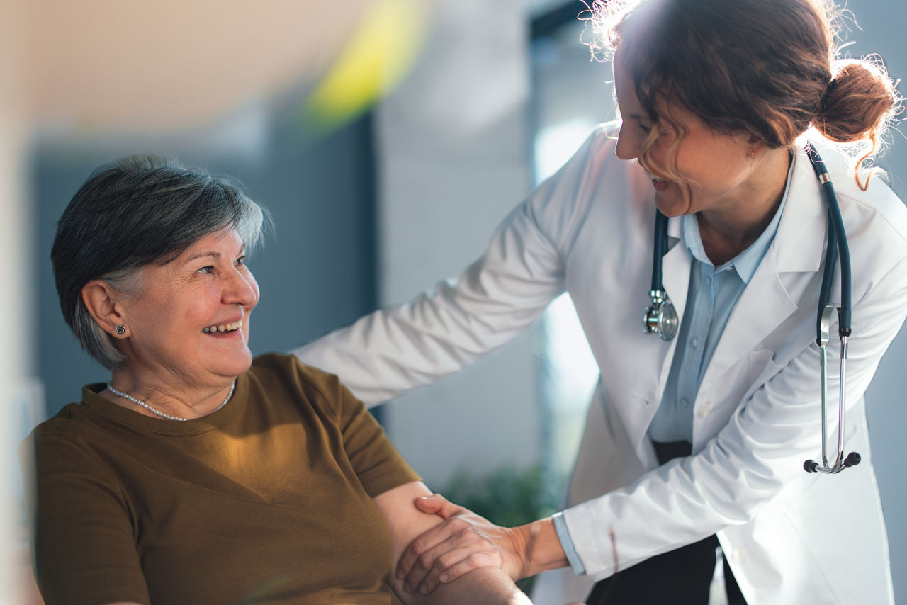 Senior woman smiles while a friendly female doctor in a white coat comforts her in a bright doctor office, conveying trust, compassionate medical care and positive patientâprovider connection.