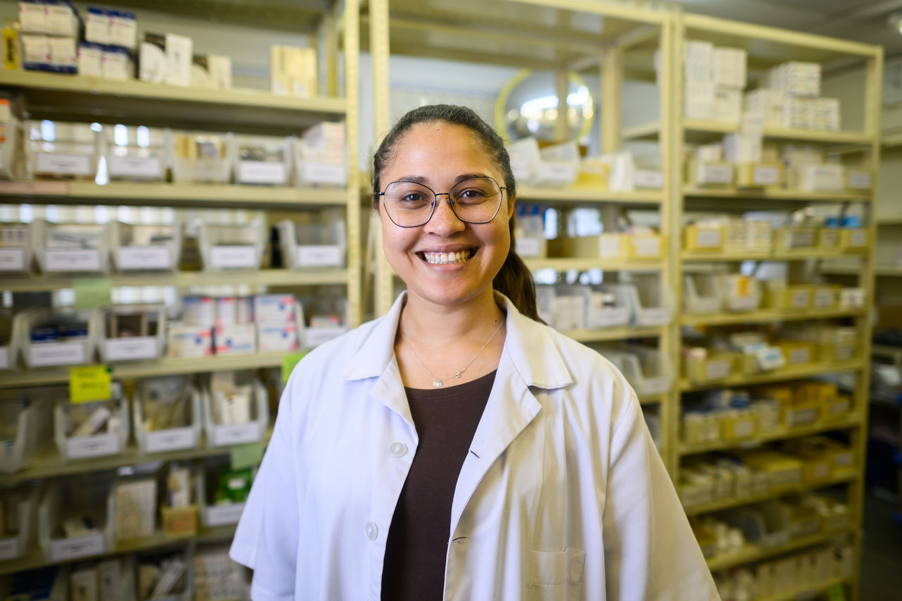 A cheerful pharmacist wearing a white lab coat stands in an oncology department pharmacy. Behind her, organized shelves containing various medical supplies and medications are visible. The scene conveys professionalism, healthcare expertise, and a welcoming atmosphere in a clinical setting. The focus highlights the efficiency and dedication within the oncology department.