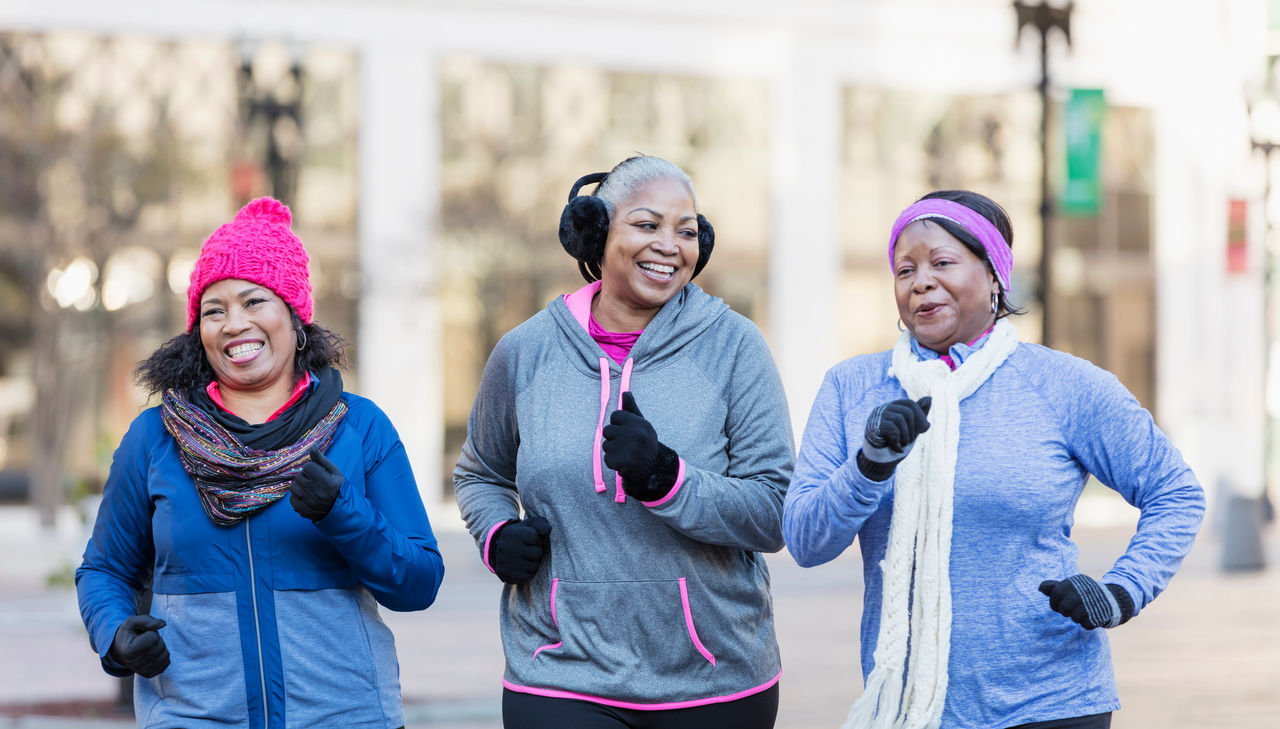 Three African-American women exercising together in the city, jogging or power walking, laughing and conversing. A building is out of focus in the background. The one on the left is in her 60s and her friends are mature women in their 40s and 50s. It is autumn or winter as they are wearing warm workout clothing, gloves, hats and ear muffs.